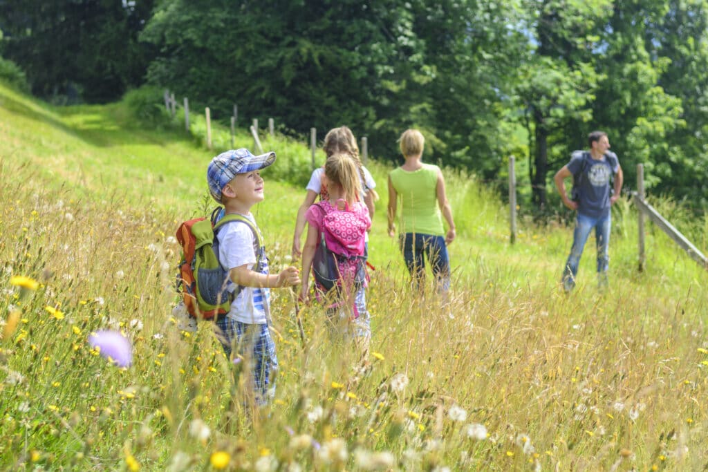 Kinder laufen mit Rucksäcken auf einer Wiese. Im Hintergrund steht ein Betreuer.