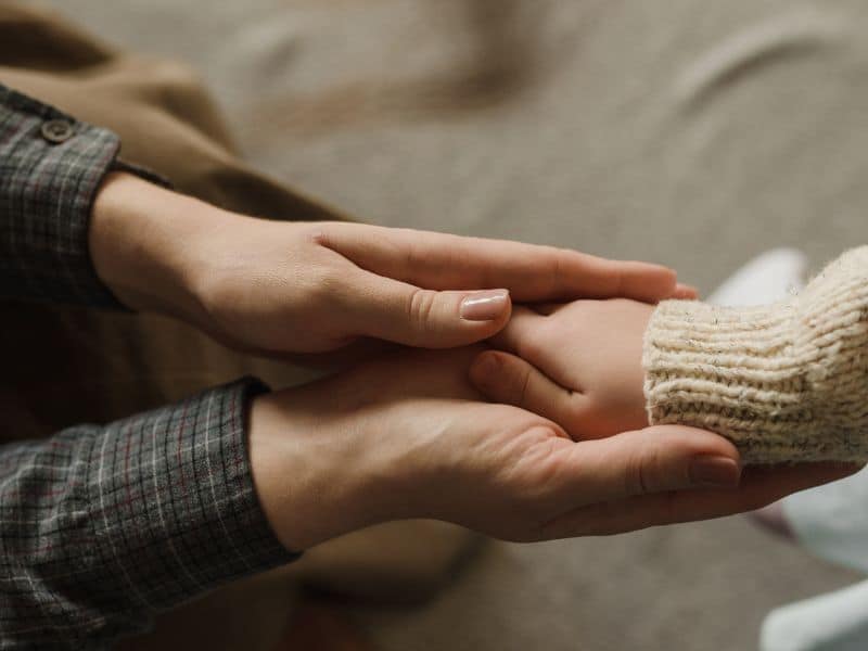 Eine erwachsene Person hält sanft die Hand eines Kindes. Beide tragen langärmlige Kleidung – die erwachsene Person ein kariertes Hemd, das Kind einen beigen Strickpullover. Die Szene vermittelt Wärme, Geborgenheit und Fürsorge.