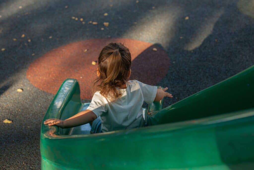 Ein kleines Kind mit braunen Haaren sitzt auf einer grünen Rutsche auf einem Spielplatz. Das Kind ist von hinten zu sehen und trägt ein weißes T-Shirt. Der Boden ist mit farbigem Fallschutzbelag bedeckt.