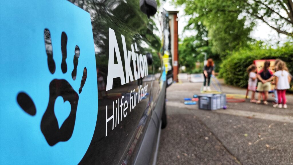 Links im Vordergrund ist die Tür eines schwarzen Transporters mit der weißen Aufschrift "Aktion Hilfe für Kinder" zu sehen. Neben der Schrift ist eine schwarze Kinderhand auf hellblauem Hintergrund zu sehen. Unscharf rechts im Bild sind mehrere Kinder zu sehen, die mit dem Rücken zur Kamera stehen.