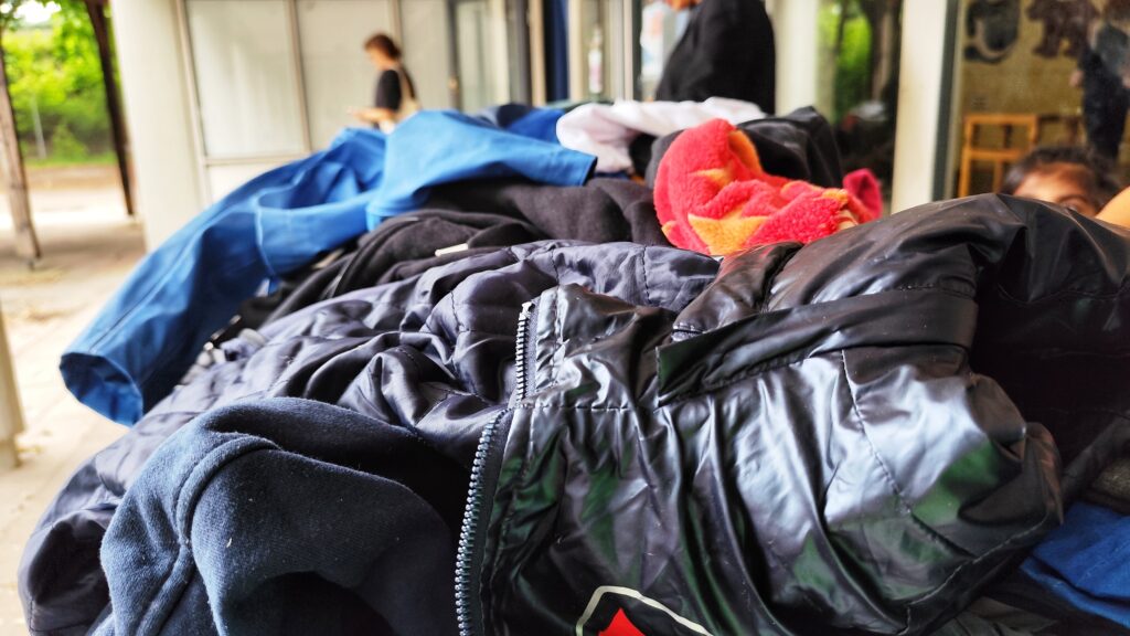 Ein großer Haufen Kleidung liegt auf einem Tisch oder einer Ablage im Vordergrund. Zu sehen sind verschiedene Jacken, Pullis und ein rotes Handtuch mit gelbem Muster. Im Hintergrund befinden sich eine Glasfassade und unscharf dargestellte Personen, darunter ein Kind, das rechts aus dem Bildrand schaut. Die Szene stellt eine Kleidungstauschbörse in einem Schulgebäude dar.