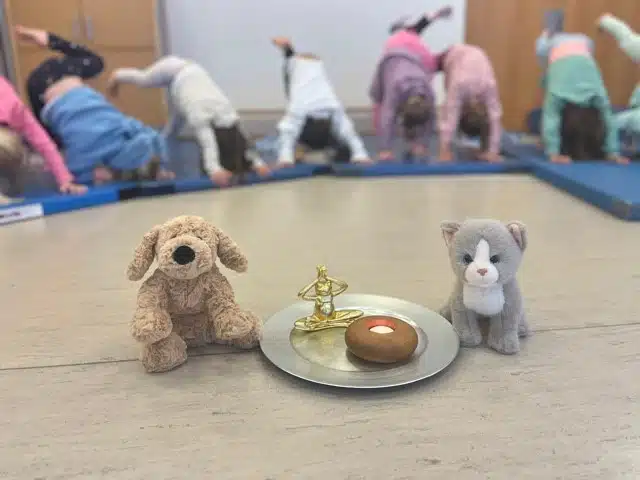 Mehrere Kinder machen eine Yoga-Übung in einem Raum und stehen dabei in der Position des herabschauenden Hundes. Im Vordergrund stehen zwei Stofftiere, ein Hund und eine Katze, neben einem Tablett mit einer kleinen Kerze und einer goldfarbenen Figur in Meditationshaltung.