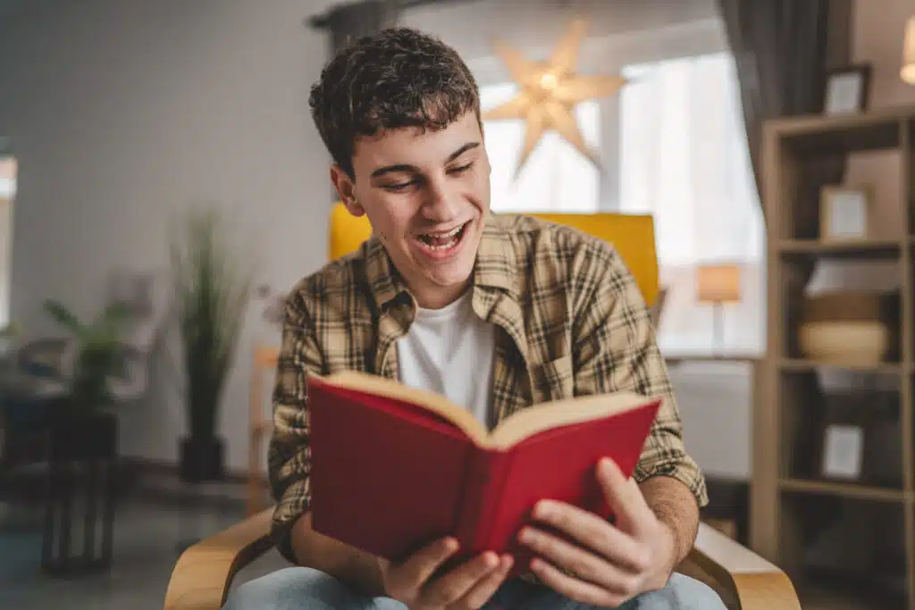 Ein junger Mann sitzt in einem gemütlichen Zimmer und liest lachend ein rotes Buch. Er trägt ein kariertes Hemd über einem weißen T-Shirt und wirkt entspannt und fröhlich. Im Hintergrund sind eine leuchtende Sternenlampe, Pflanzen und Regale mit Büchern zu sehen.
