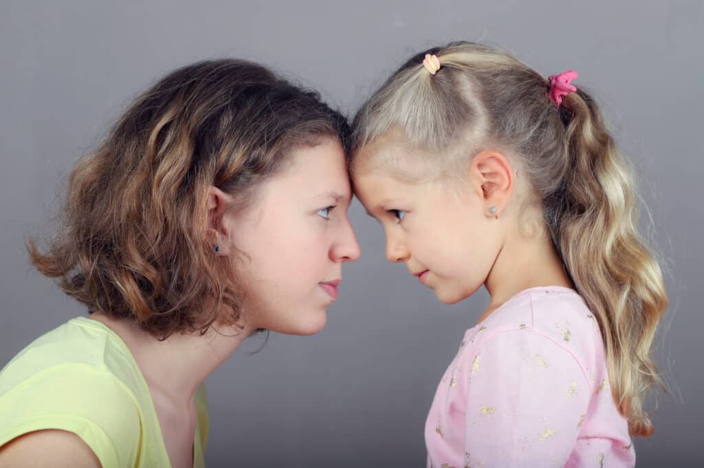 Eine Mutter und ihrer jungen Tochter stehen sich gegenüber und sie lehnen ihre Stirnen aneinander. Die Mutter hat schulterlanges braunes Haar mit Strähnchen und trägt ein hellgrünes Oberteil. Das kleine Mädchen hat blonde Haare, die zu Zöpfen gebunden sind mit bunten Haargummis, und trägt ein rosa Oberteil mit kleinen Mustern. Beide sind im Profil zu sehen und schaffen eine intime, liebevolle Verbindung, während sie sich vor einem neutralen grauen Hintergrund anblicken.