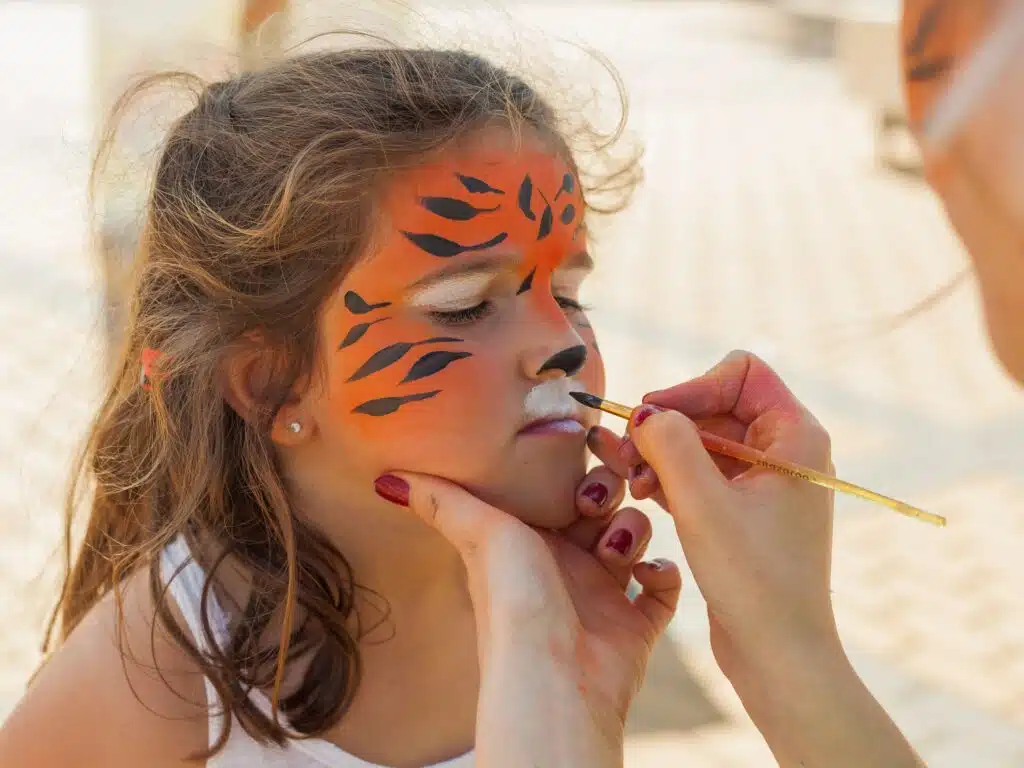 Ein Kind mit geschlossenen Augen bekommt ein Gesichtsmakeup. Eine Person malt mit einem Pinsel Tigerstreifen und weiße Akzente auf das Gesicht des Kindes.