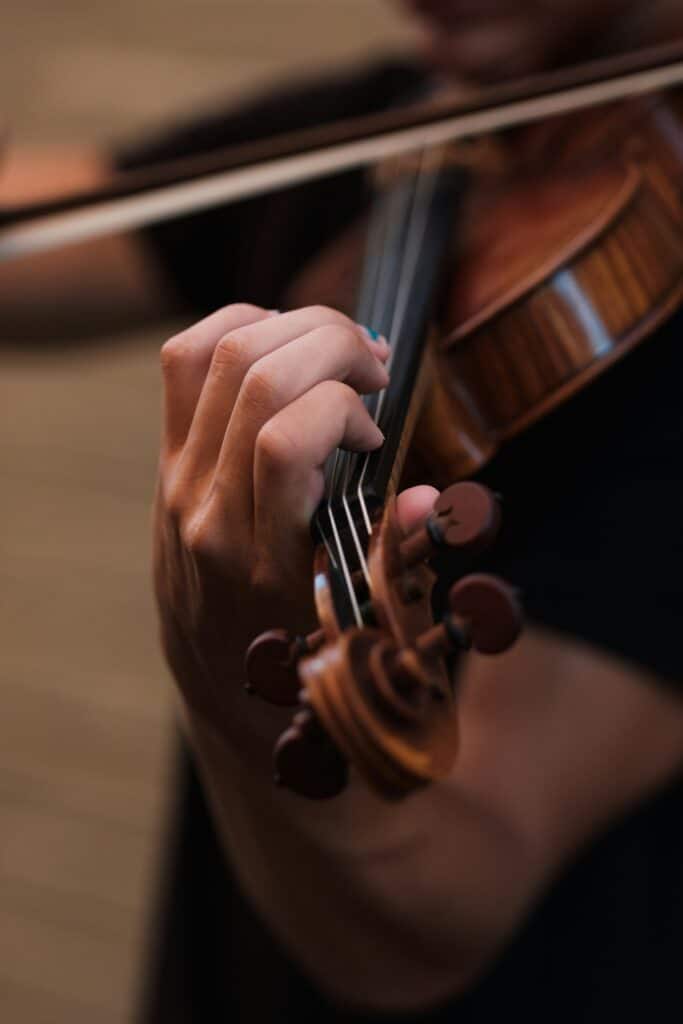 Nahaufnahme einer Person, die eine Geige spielt. Die linke Hand greift die Saiten am Griffbrett, während der Bogen über die Saiten geführt wird. Der Hintergrund ist unscharf, der Fokus liegt auf der Hand und dem Instrument.