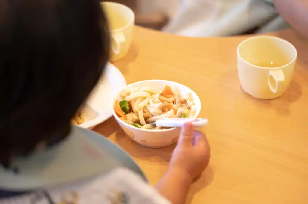 Ein kleines Kind sitzt an einem Holztisch und hält einen Löffel in der Hand. Vor ihm steht eine Schüssel mit Udon-Nudeln, Gemüse und etwas Brühe. Neben der Schüssel stehen zwei gelbe Becher, einer davon enthält ein Getränk.