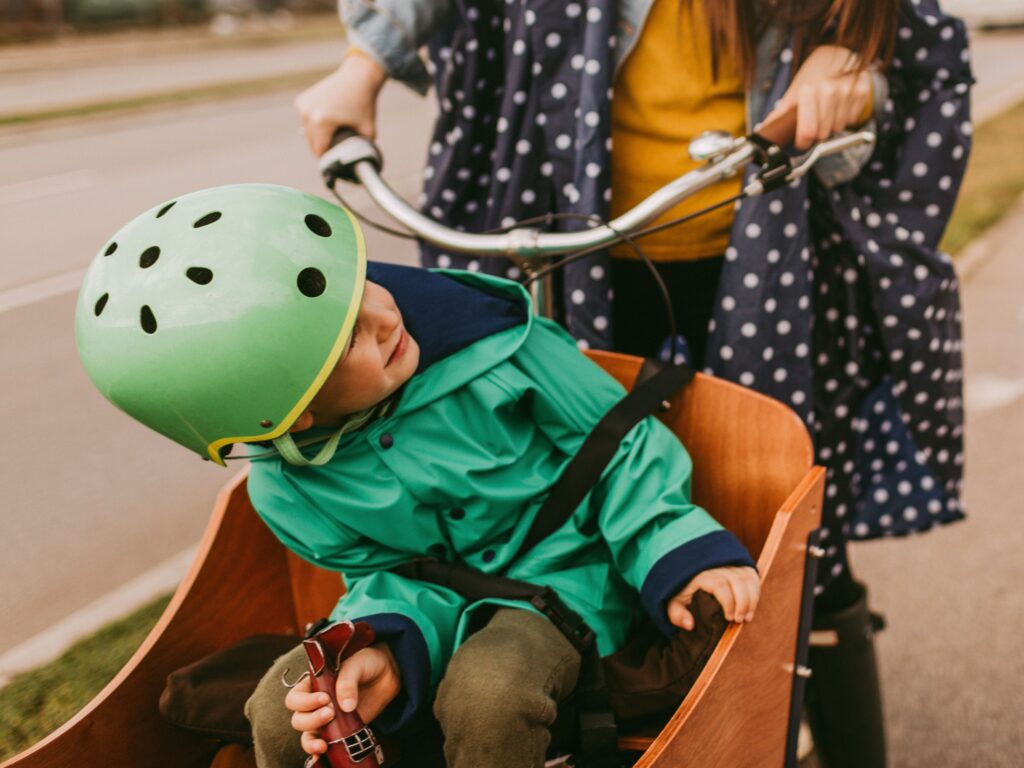 Ein Kind mit grünem Helm und grüner Regenjacke sitzt angeschnallt in einer hölzernen Fahrradkiste. Eine erwachsene Person, die eine gepunktete Regenjacke trägt, hält den Lenker des Fahrrads. Die Szene spielt auf einer Straße oder einem Radweg.