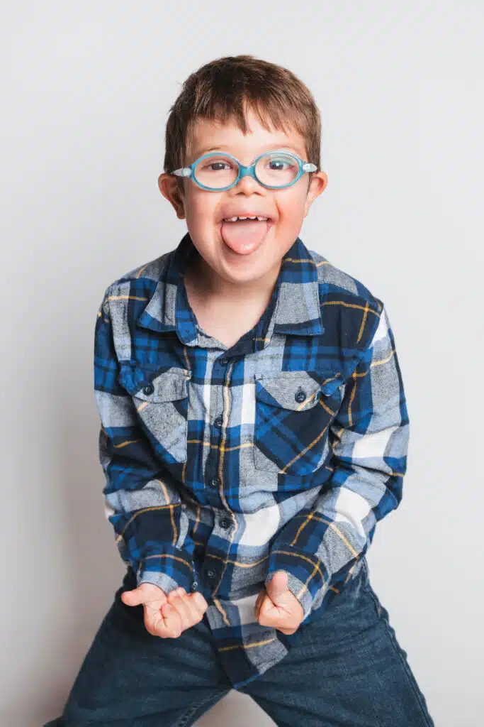 Ein lachendes Kind mit kurzen braunen Haaren und runder blauer Brille streckt fröhlich die Zunge heraus. Es trägt ein blau kariertes Hemd und steht vor einem hellen, neutralen Hintergrund mit geballten Fäusten in einer spielerischen Pose.