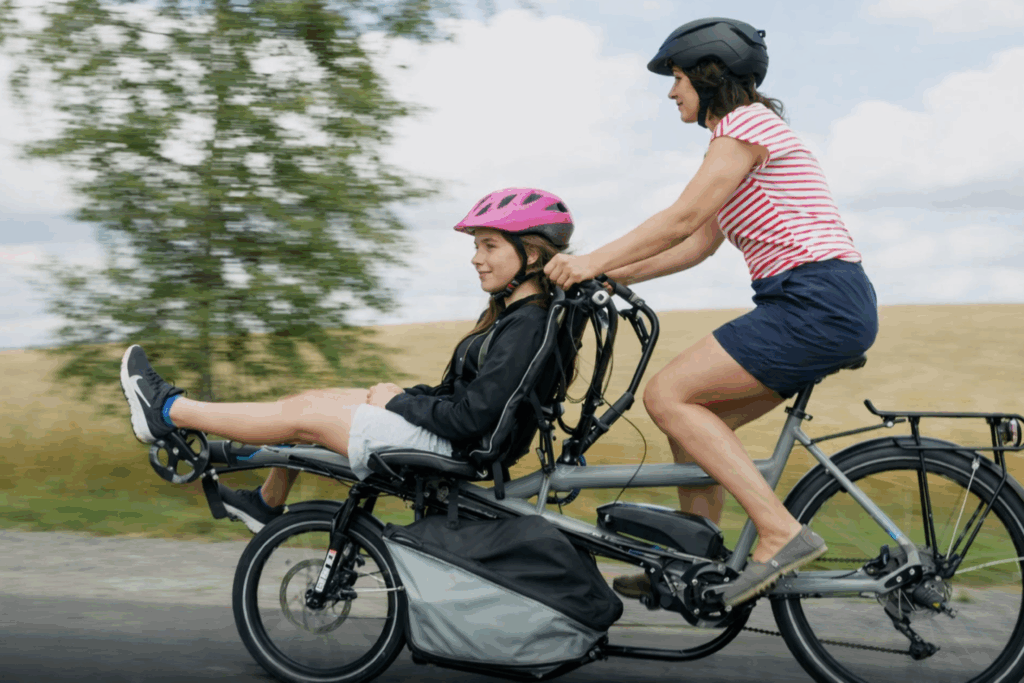 Eine Frau fährt mit ihrer Tochter auf einem grauen Lastenfahrrad durch eine Landschaft. Das Kind sitzt vorne im Transportsitz, beide tragen Fahrradhelme und lachen.