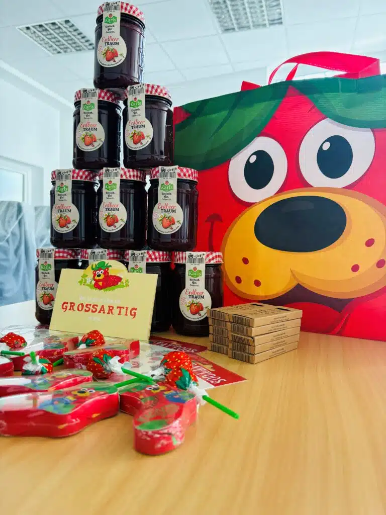 Stapel aus Erdbeer-Fruchtaufstrich-Gläsern pyramidenförmig auf einem Tisch arrangiert, daneben eine rote Geschenktasche mit Cartoon-Hundegesicht, ein „Grossartig“-Schild sowie verpackte Süßigkeiten und kleine Schokoladentafeln im Vordergrund.