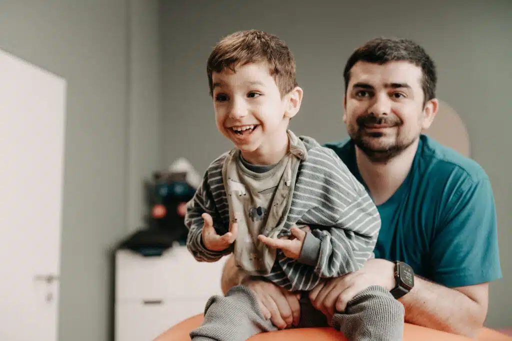Ein lächelnder Junge sitzt während einer Therapiesitzung auf einem Gymnastikball und strahlt. Hinter ihm ist der Physiotherapeut.