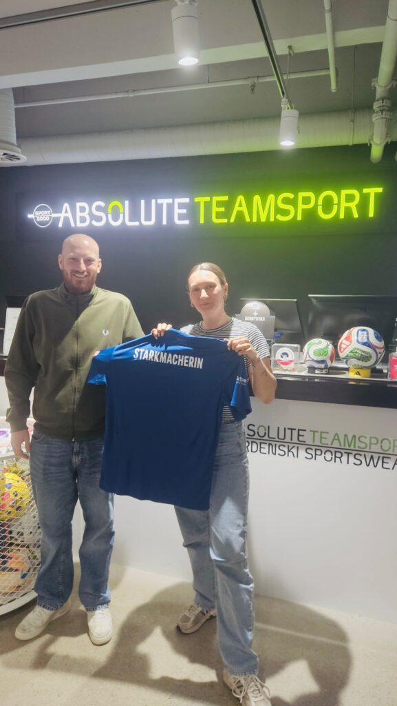 Zwei Personen, ein Mann mit Glatze und Vollbart und eine Frau mit blonden Haaren, stehen in einem Sportgeschäft und halten gemeinsam ein blaues Trikot hoch. Auf der Rückseite des Trikots steht in weißen Buchstaben „STARKMACHERIN“. Im Hintergrund sind mehrere Fußbälle und ein Verkaufstresen zu sehen. Zudem steht auf der Wand "ABSOLUTE TEAMSPORT".