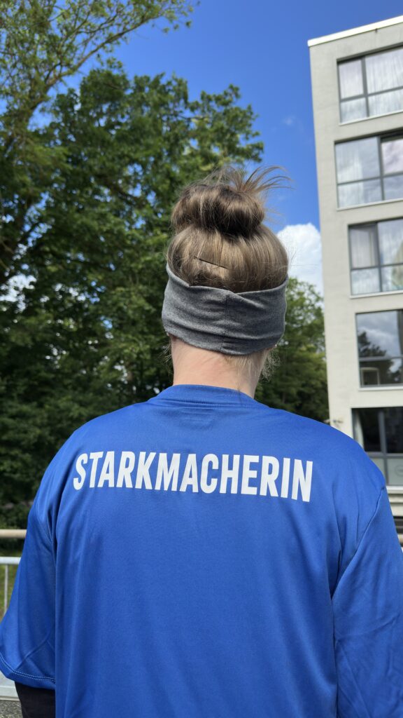Eine Person steht draußen vor einer grünen Baumkulisse und einem modernen Gebäude. Sie ist von hinten zu sehen, die Haare zu einem Dutt gebunden und mit einem grauen Stirnband gehalten. Auf dem blauen T-Shirt steht in großen weißen Buchstaben „STARKMACHERIN“. Der Himmel ist blau und die Szene wirkt hell und freundlich.