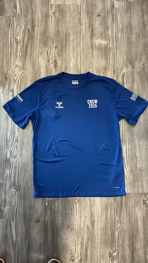 Ein blaues T-Shirt liegt ausgebreitet auf einem hellen Holzfußboden. Auf der Vorderseite befinden sich ein Herstellerlogo und der weiße Schriftzug „CREW 2026“ im Brustbereich. Zusätzlich sind auf beiden Ärmeln weiße Aufdrucke zu sehen. Das Foto zeigt das Shirt vollständig von oben und hebt Design, Farbe und Platzierung der Druckelemente hervor.