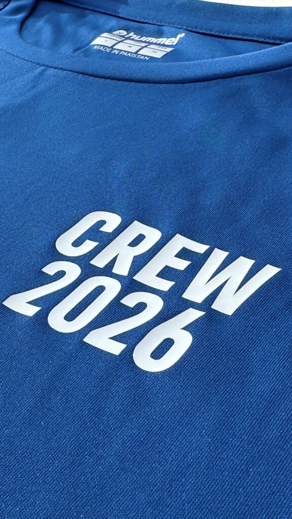 Das Bild zeigt eine Detailaufnahme eines blauen T-Shirts. Im Mittelpunkt steht der große weiße Aufdruck „CREW 2026“, der schräg über die Vorderseite verläuft. Am Kragen ist das Herstellerlogo von Hummel sichtbar. Die Aufnahme hebt die Farbe, den Stoff und die Bedruckung des Shirts hervor.