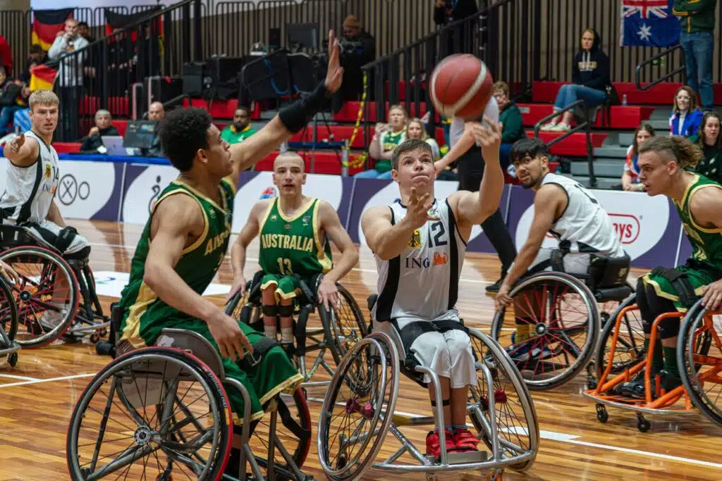 Rollstuhlbasketballspieler der deutschen und australischen Mannschaft kämpfen während eines Spiels um den Ball. Ein deutscher Spieler im weißen Trikot mit der Nummer 12 wirft den Ball auf den Korb, während mehrere australische Gegenspieler in grünen Trikots ihn zu blockieren versuchen; im Hintergrund verfolgen Zuschauerinnen und Zuschauer die Partie.