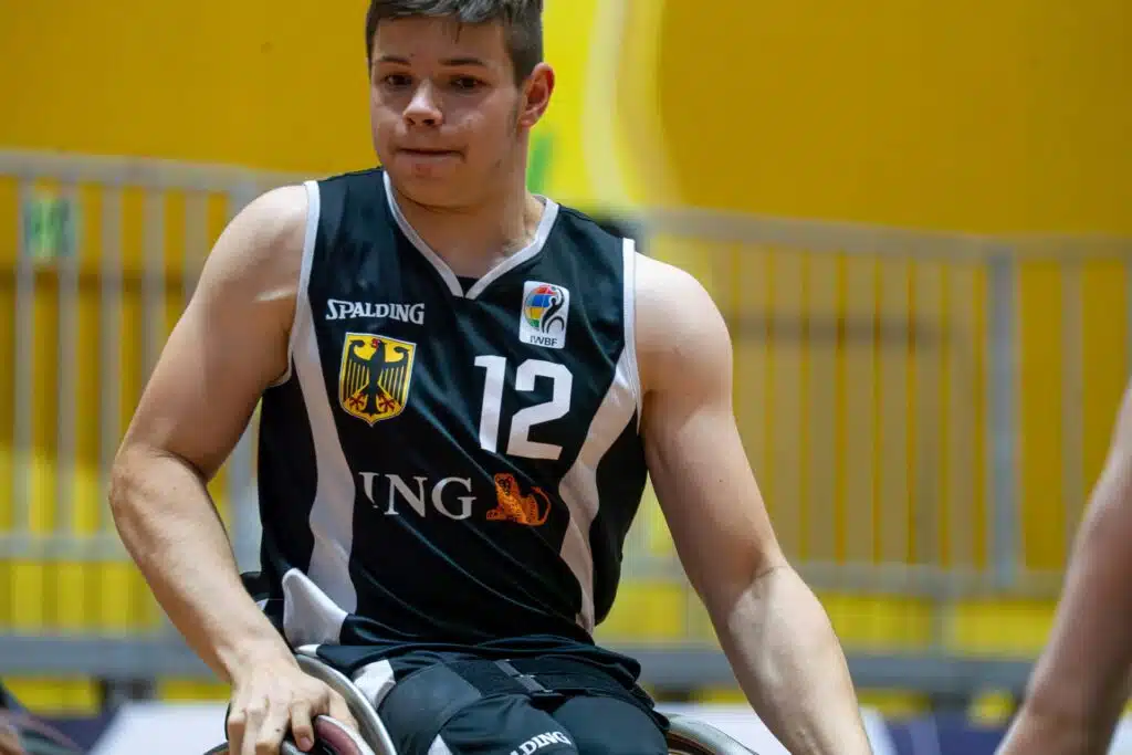 Nahaufnahme eines Rollstuhlbasketballspielers im schwarzen deutschen Nationaltrikot mit der Nummer 12 und dem Logo des internationalen Rollstuhlbasketballverbands. Der Spieler sitzt in seinem Sportrollstuhl und blickt konzentriert nach vorn; im Hintergrund sind eine gelbe Hallenwand und ein Geländer zu sehen.